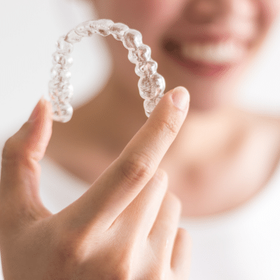 Why invisalign® might be better than traditional braces 10 Woman holding up invisalign aligner in the foreground while her face is blurred out in the background, representing why it may be better than traditional braces